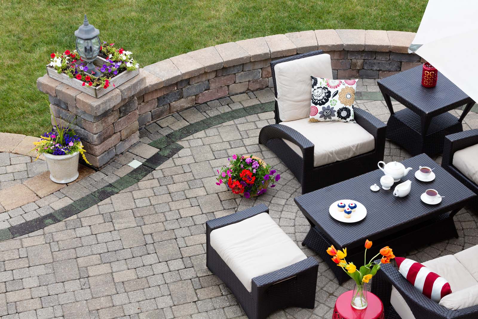 Brick paved patio with comfortable patio furniture with modern armchairs and a stool around a table set with tea and cookies alongside a low curving wall overlooking a green lawn high angle view