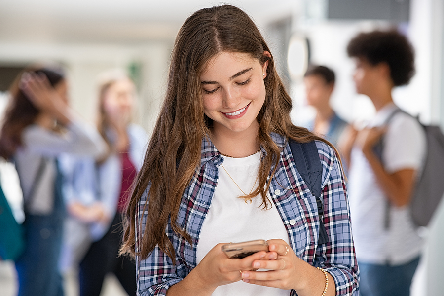 Female college student wearing a backpack smiling while looking at her phone. There are a group of students behind her but they are out of focus.