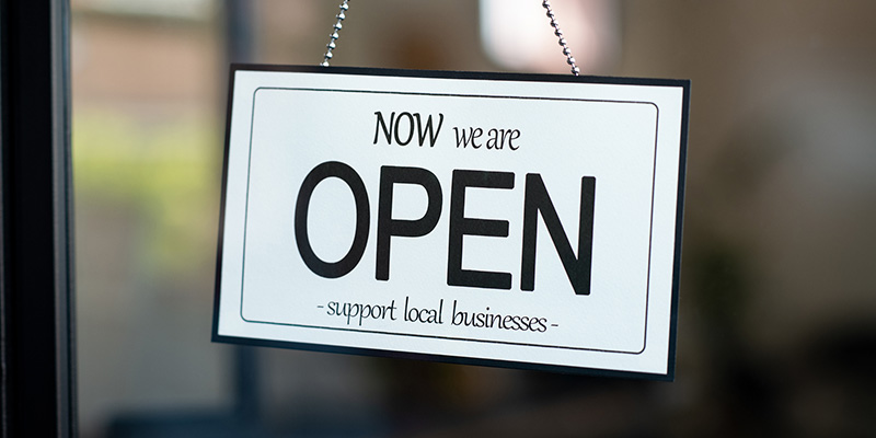 Image of a Now We Are Open sign hanging in the window of a storefront.